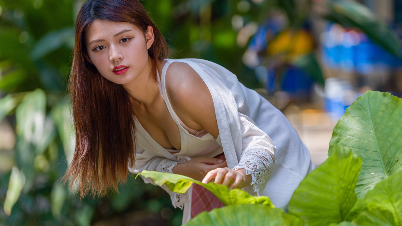 Девушка азиатка стоит у куста с зелеными листьями 