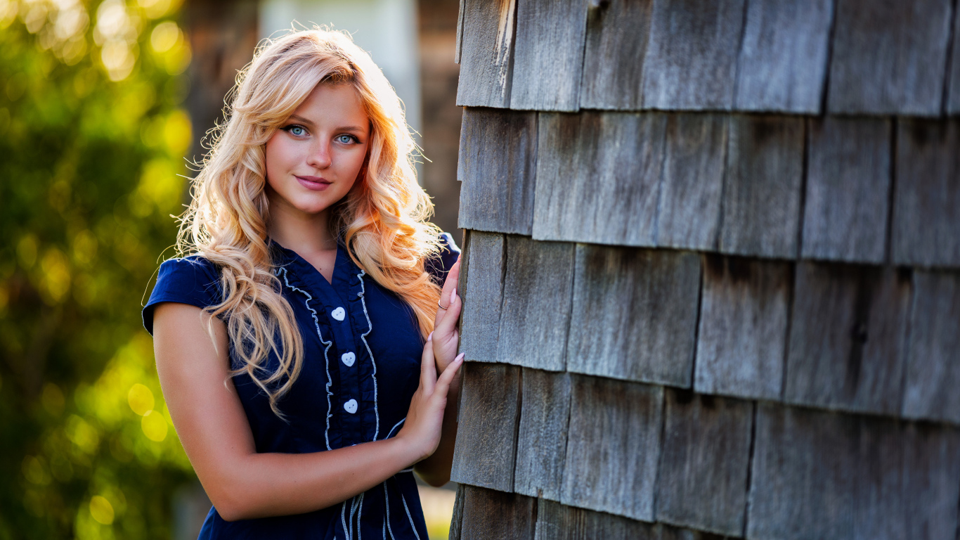 Голубоглазая блондинка в платье стоит у стены 