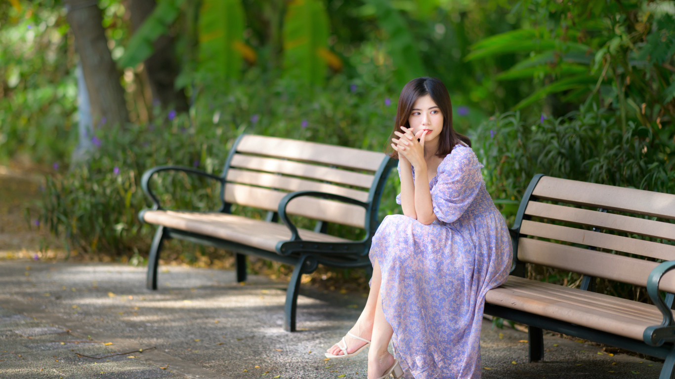 Грустная девушка азиатка в платье на лавке в парке 