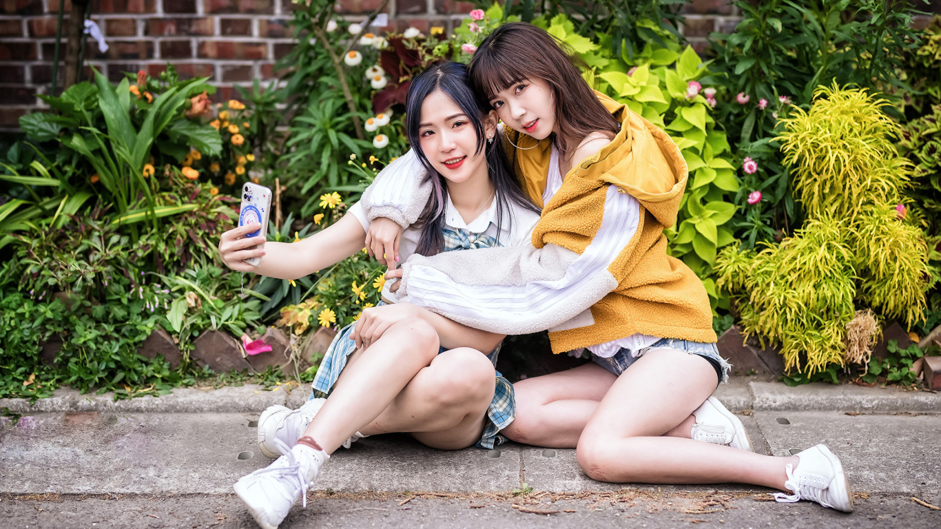 Две девушки азиатки сидят у стены с цветами 