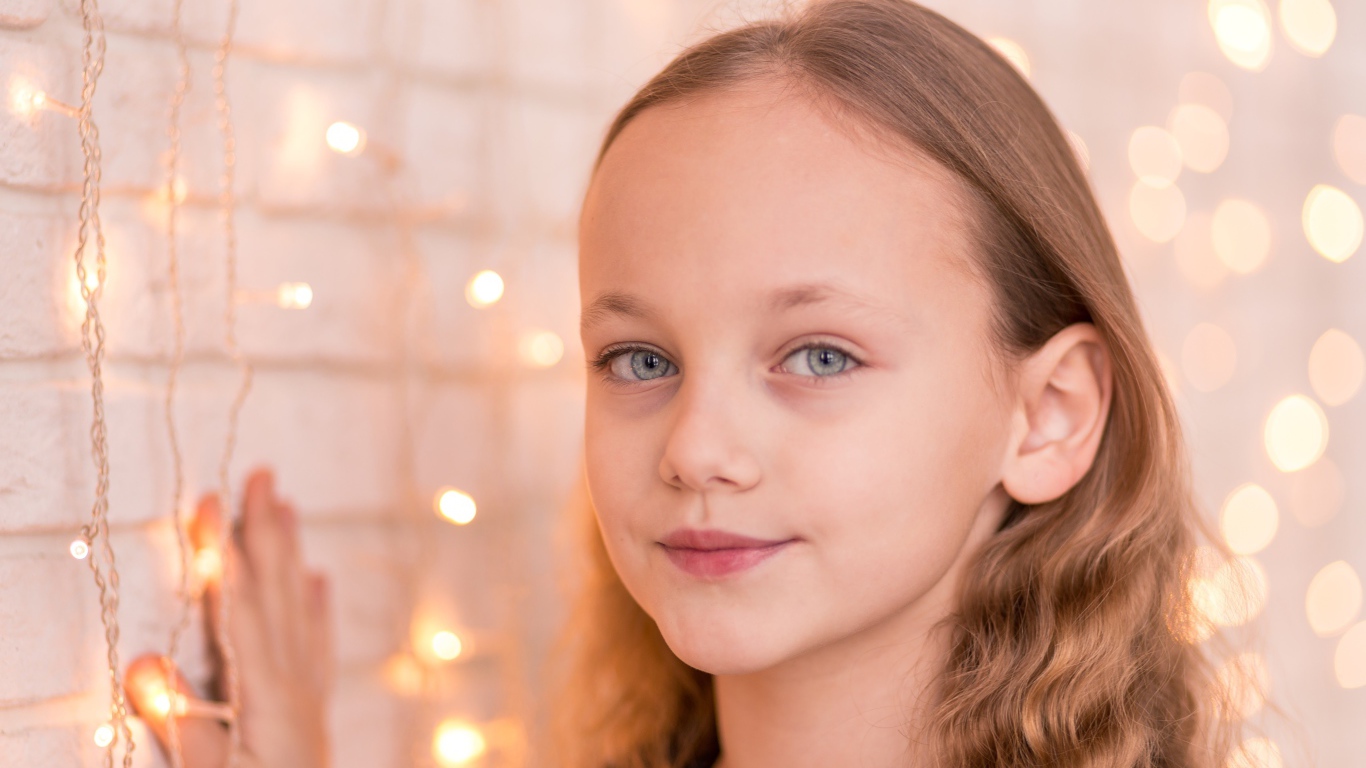 Красивая голубоглазая девочка стоит у стены с гирляндой