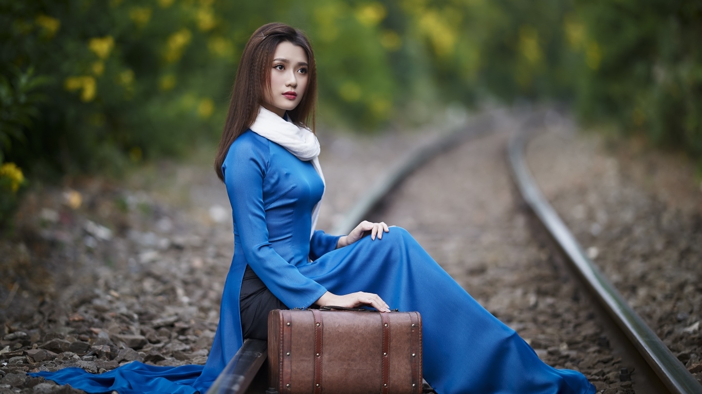 Красивая девушка азиатка сидит на рельсах с чемоданом