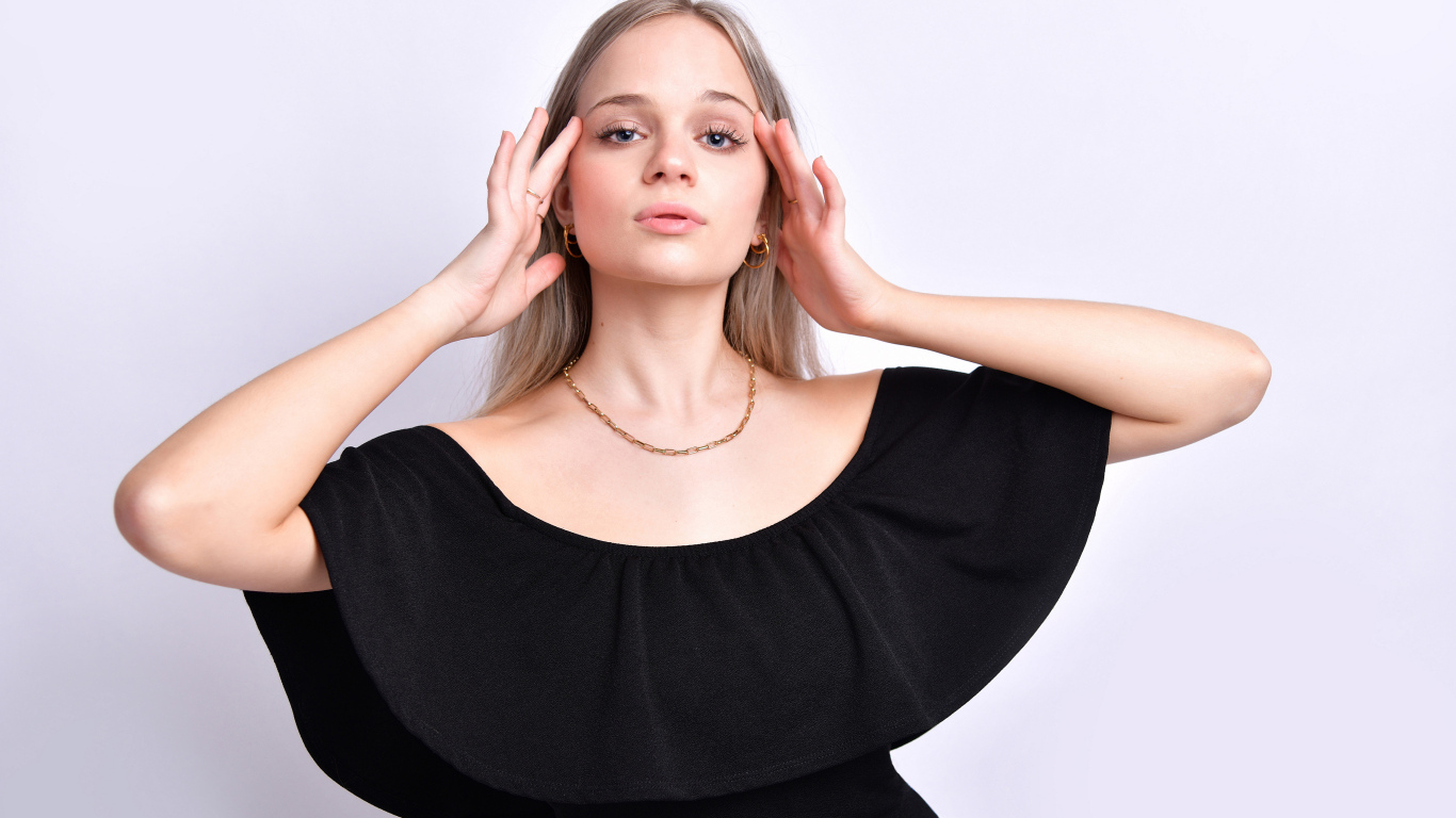 Девушка в черном платье с руками у головы на белом фоне