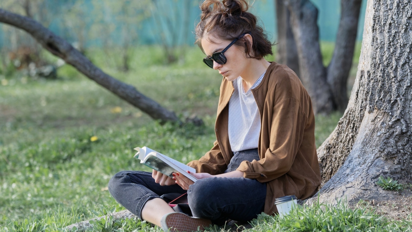 Молодая девушка сидит под деревом с книгой