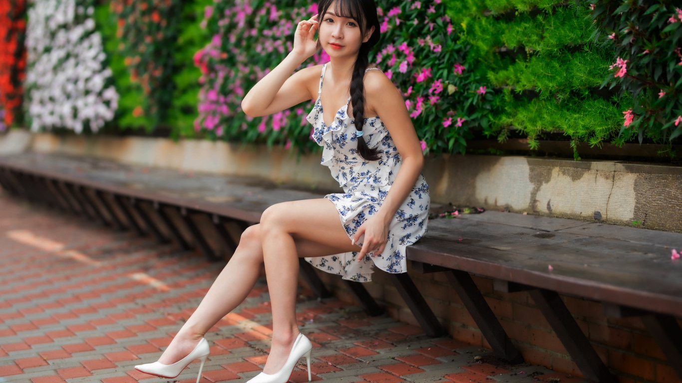 Азиатка в платье сидит на лавке у цветущих растений
