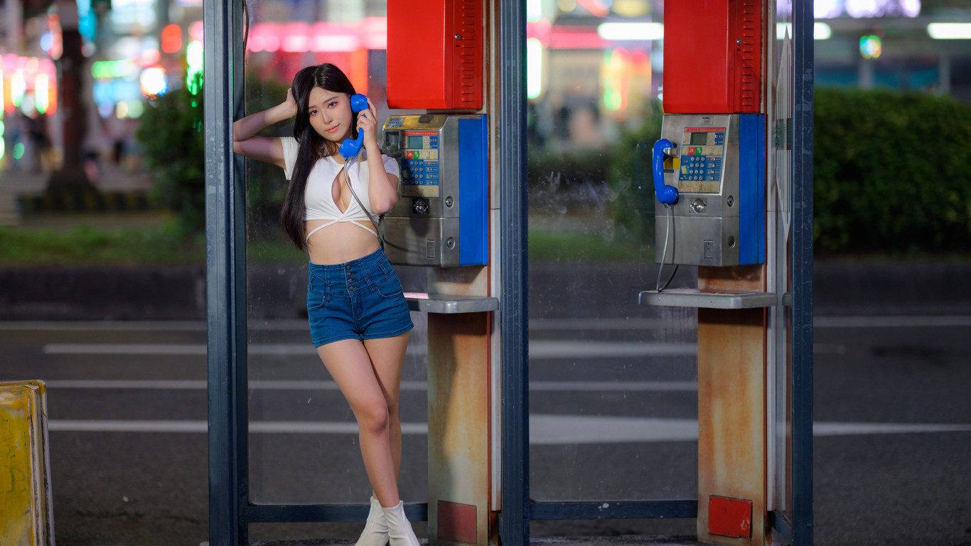 Азиатка в коротких шортах стоит у телефона-автомата