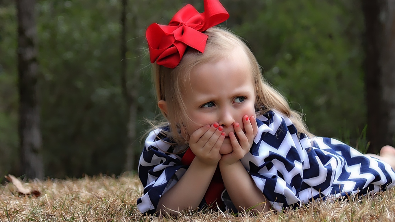 Маленькая девочка с красным бантом на голове лежит на траве