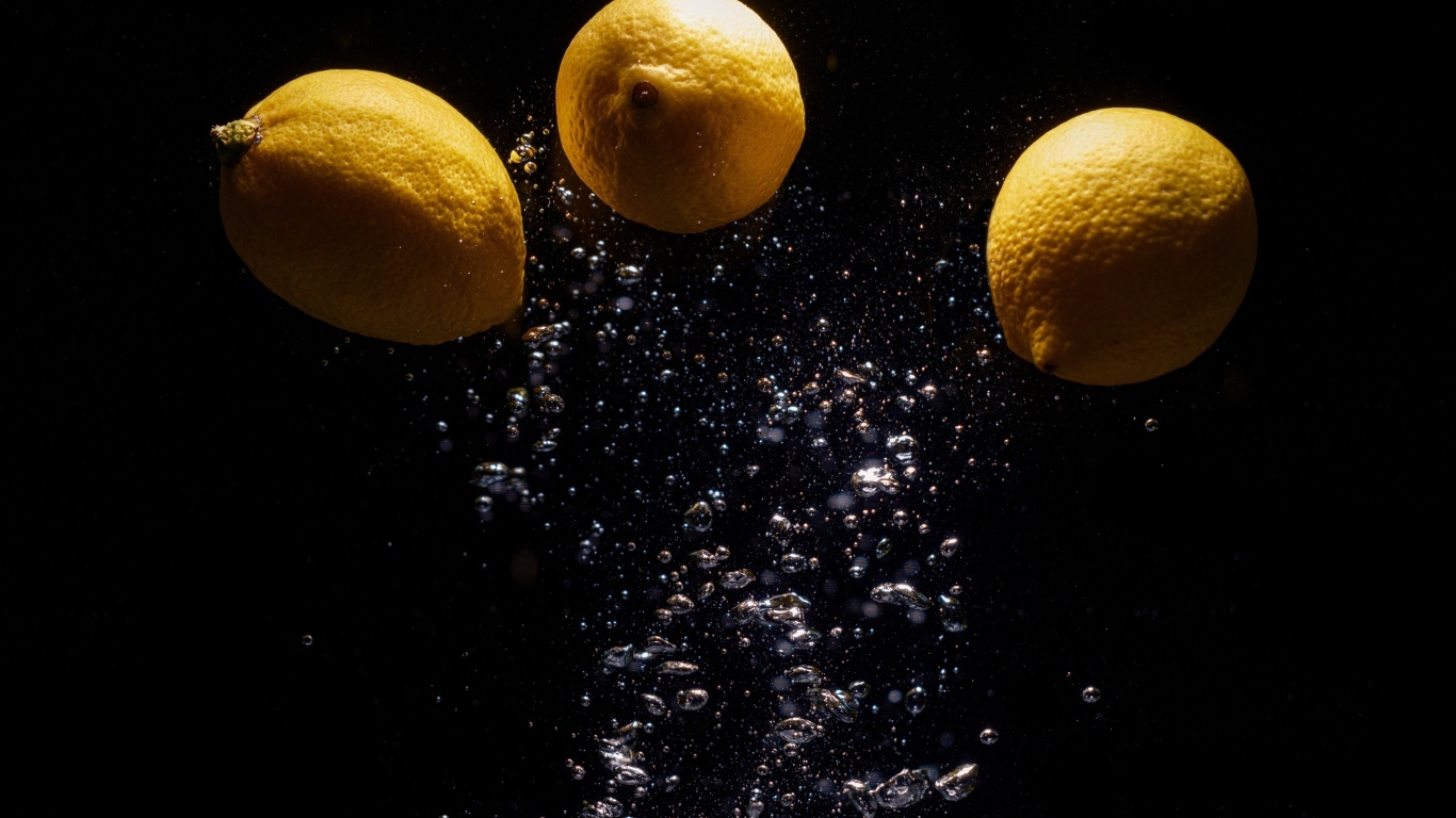 Три желтых лимона с пузырями в воде на черном фоне