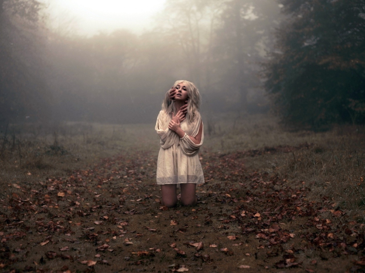 Блондинка в белом платье стоит на коленях среди леса