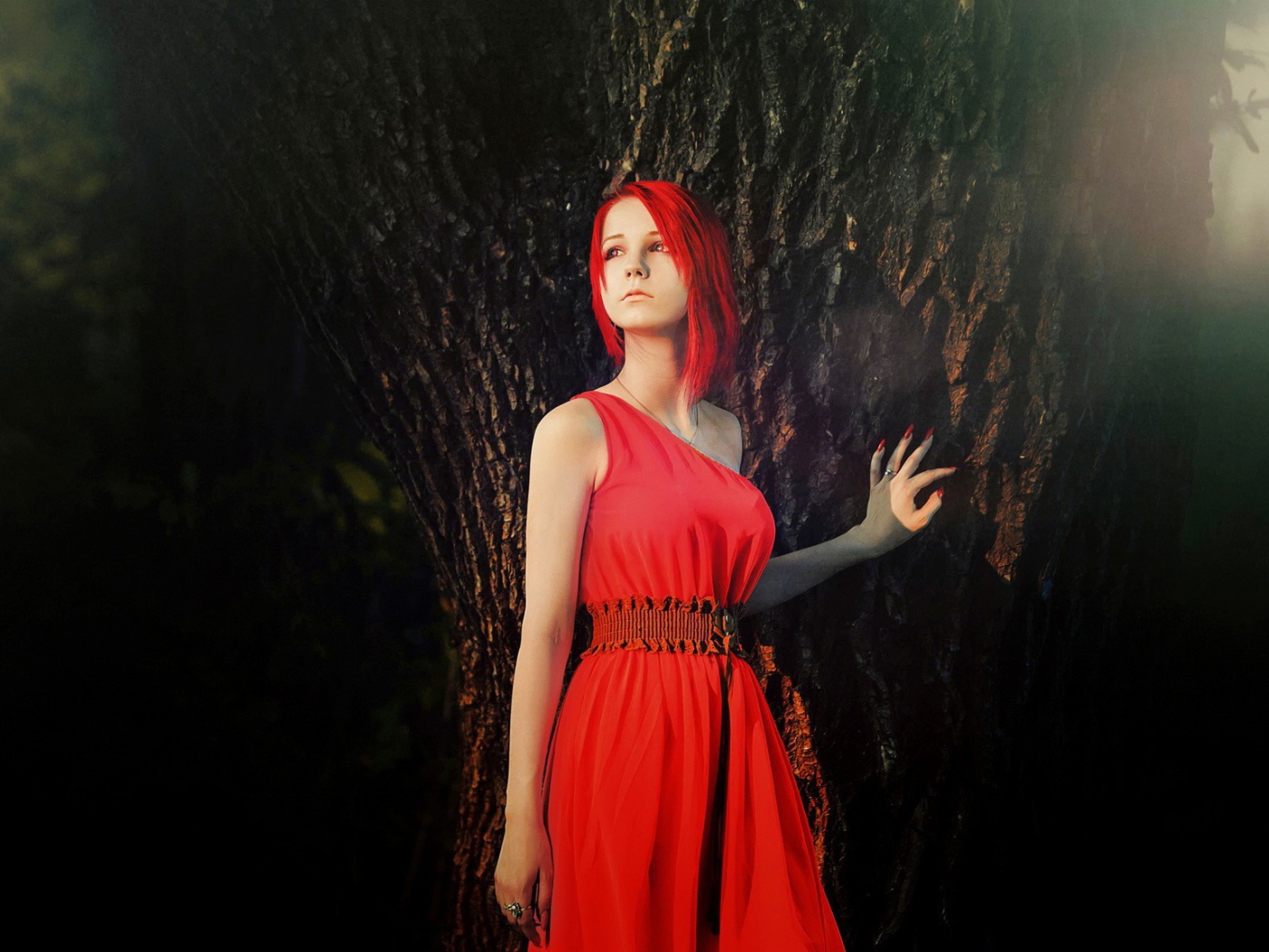 Рыжая девушка в красном платье стоит у дерева