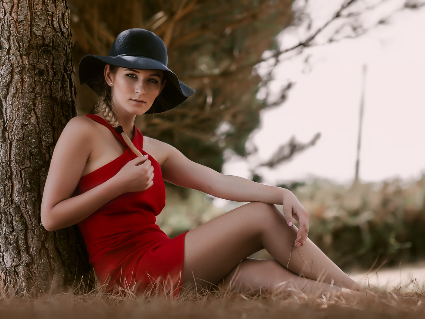 Красивая девушка в красном платье и черной шляпе сидит под деревом