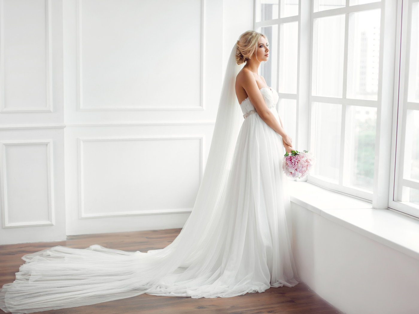 Красивая девушка в белом свадебном платье с букетом стоит у окна