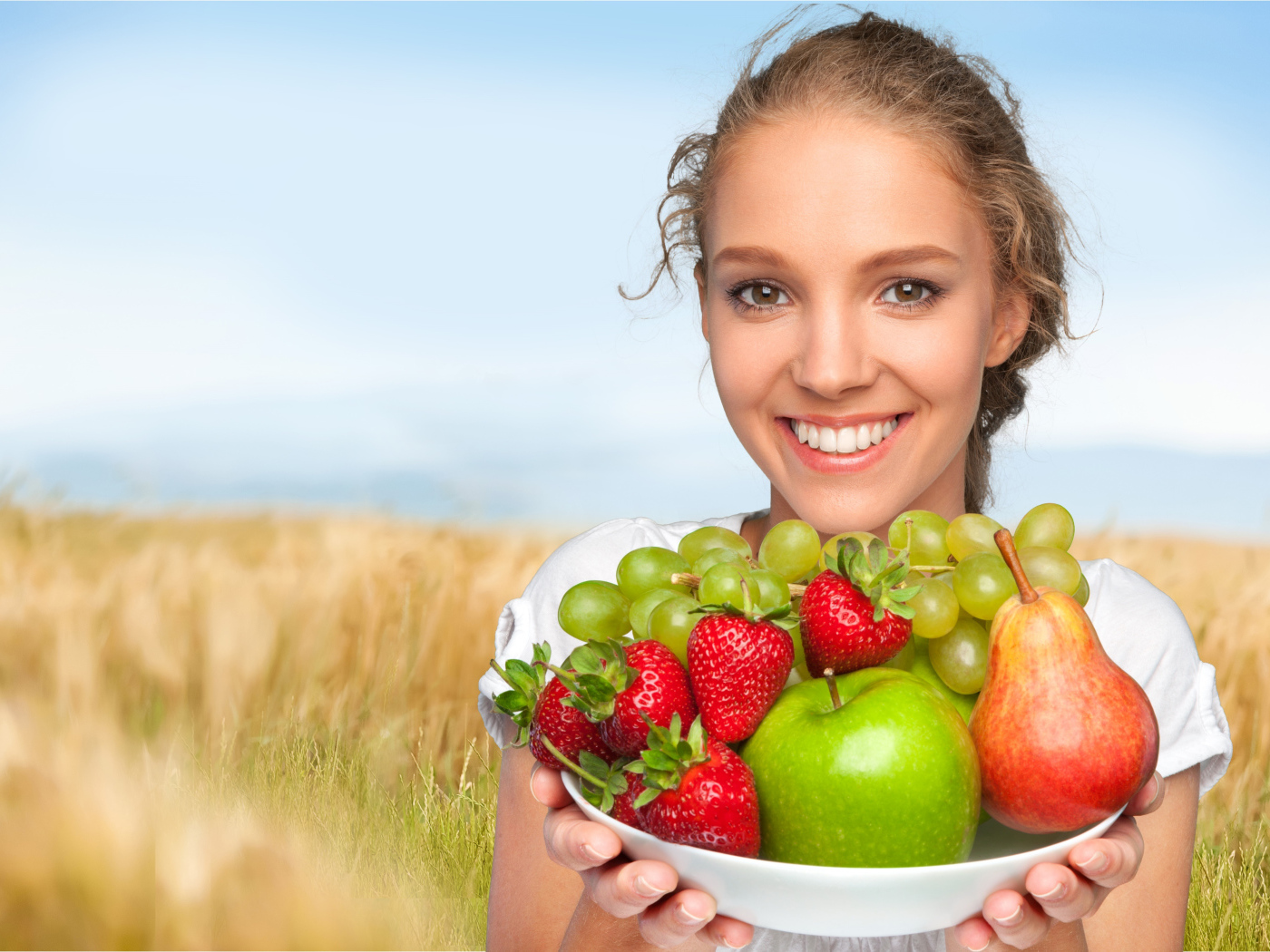Улыбающаяся девушка шатенка с тарелкой с яблоками, виноградом, клубникой и грушей в руках