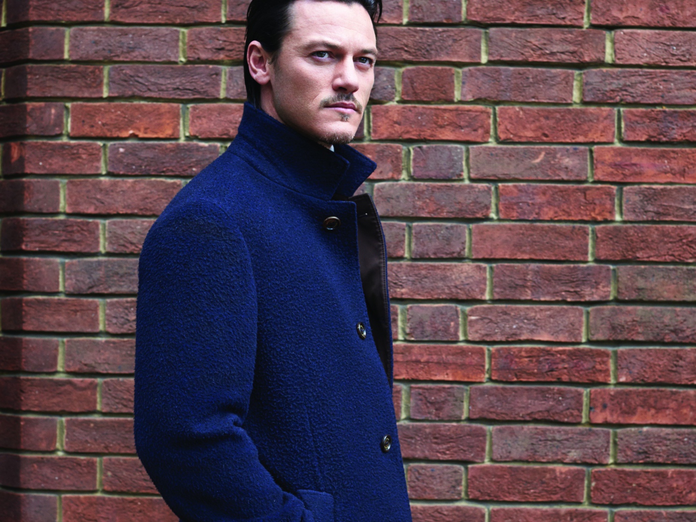 Актер Люк Эванс в синем плаще у кирпичной стены