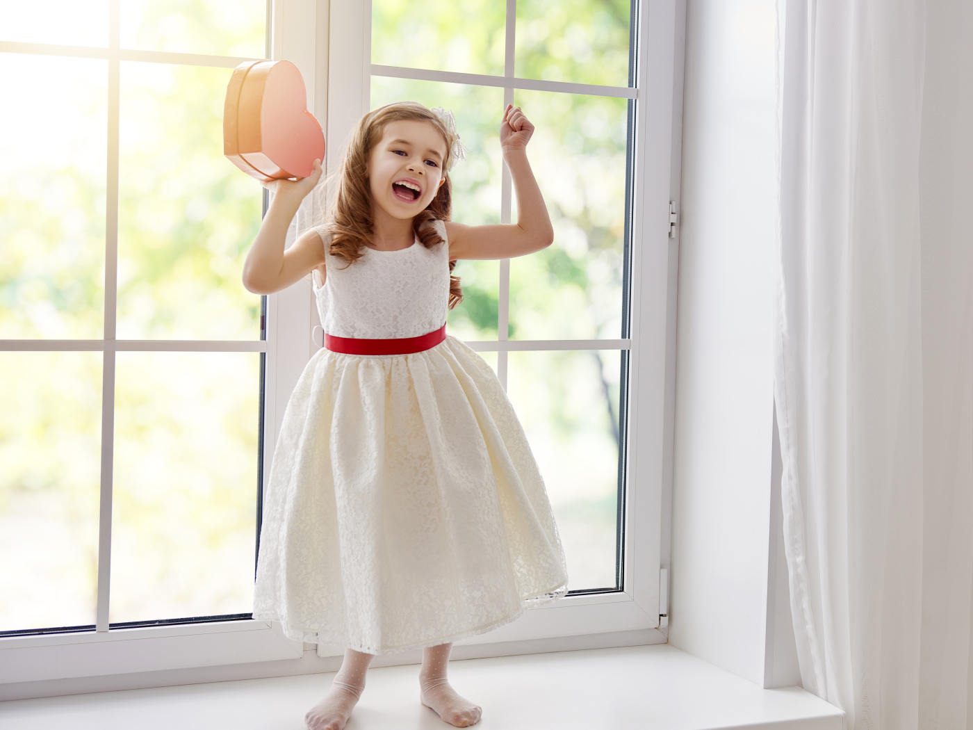 Маленькая девочка с коробкой в форме сердца в руках стоит на подоконнике
