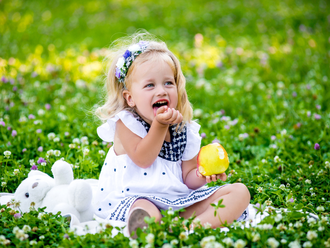 Маленькая улыбающаяся девочка с яблоком сидит на зеленой траве