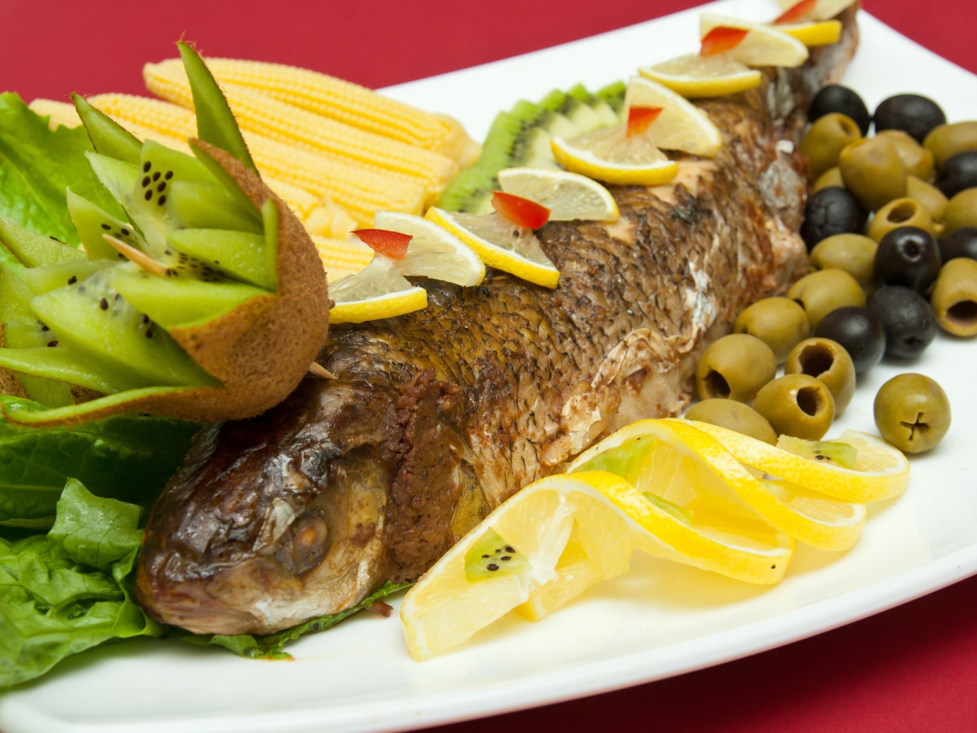 Запеченная рыба с оливками, лимоном, киви, кукурузой и листьями салата на белой тарелке