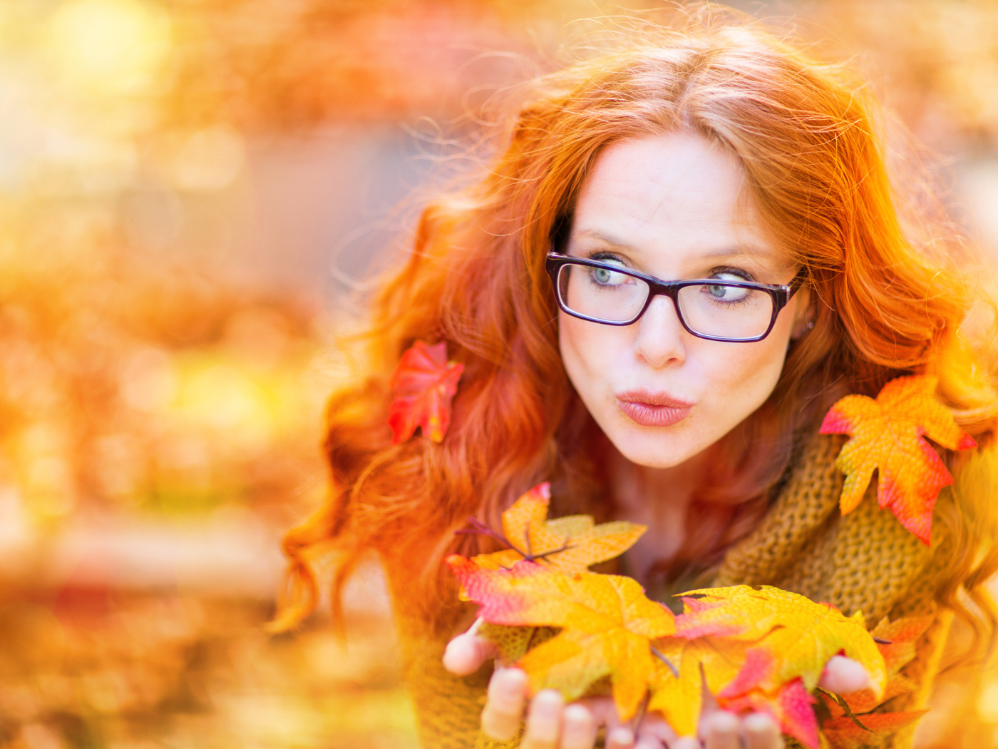 Рыжеволосая девушка в очках держит в руках опавшие листья 