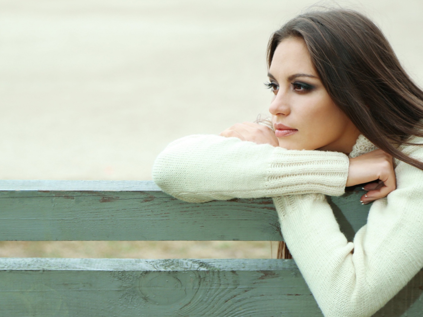 Задумчивая девушка брюнетка в белом свитере сидит на лавке