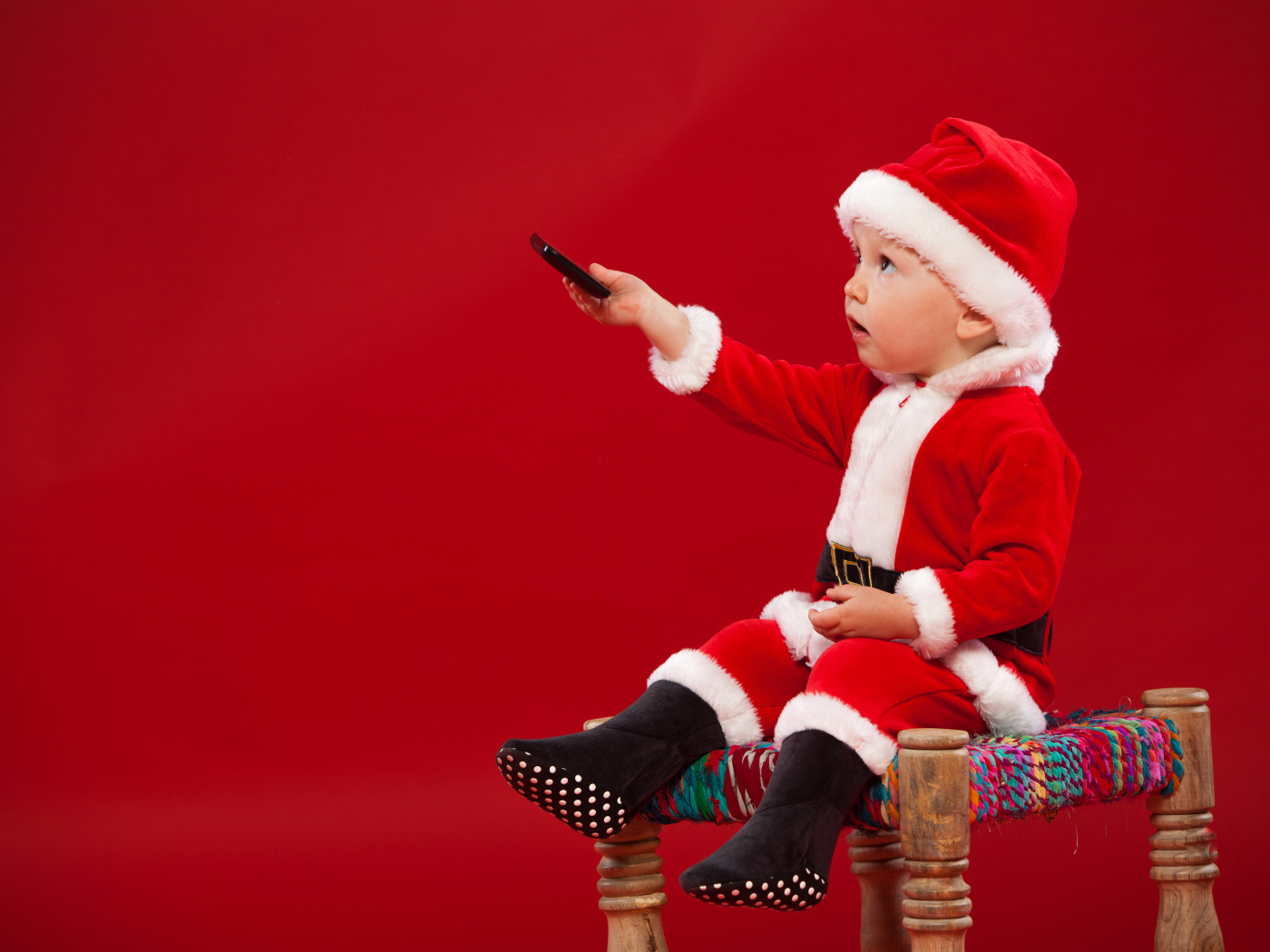 Маленький мальчик в костюме Санта Клауса сидит на стуле на красном фоне