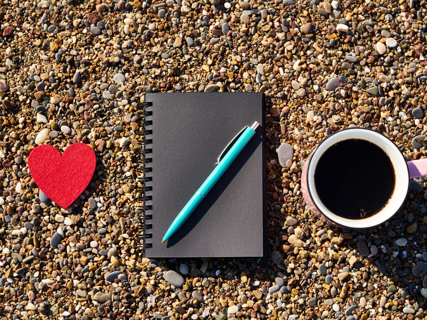 Блокнот с ручкой лет на мелкой гальке с чашкой кофе и красным сердцем 