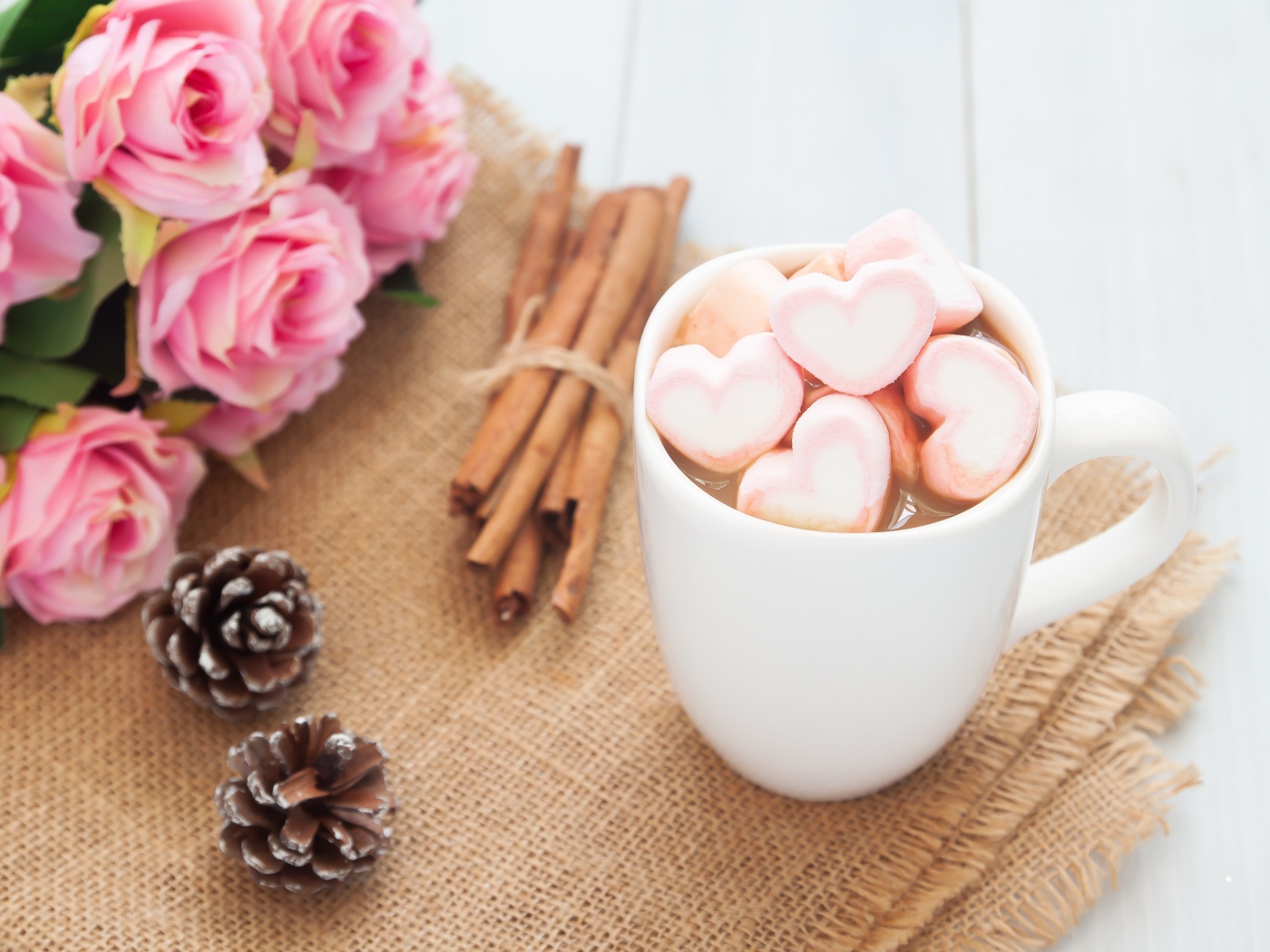 Чашка какао с маршмеллоу на столе с корицей, шишками и букетом розовых роз