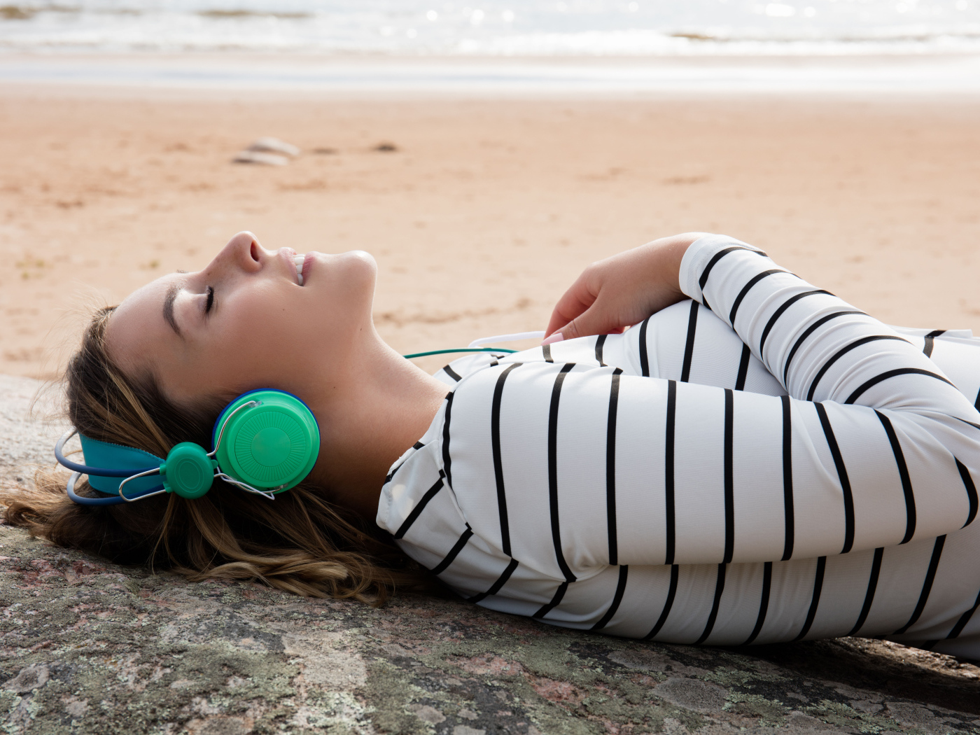 Молодая девушка лежит в наушниках на земле у моря