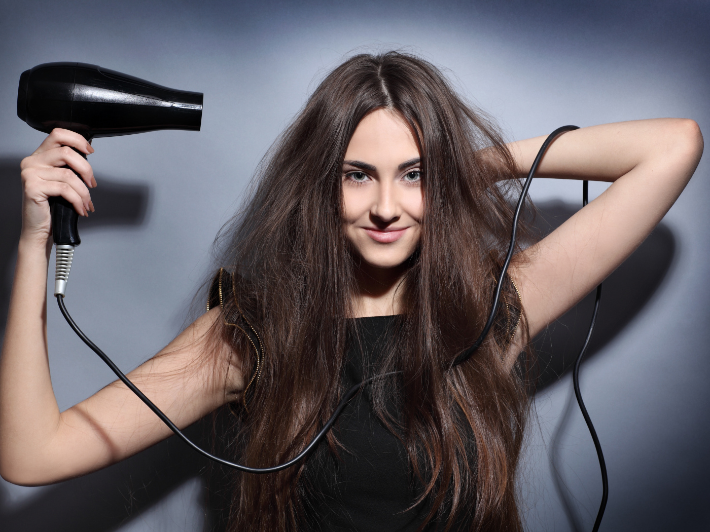 Длинноволосая девушка сушит волосы феном на сером фоне