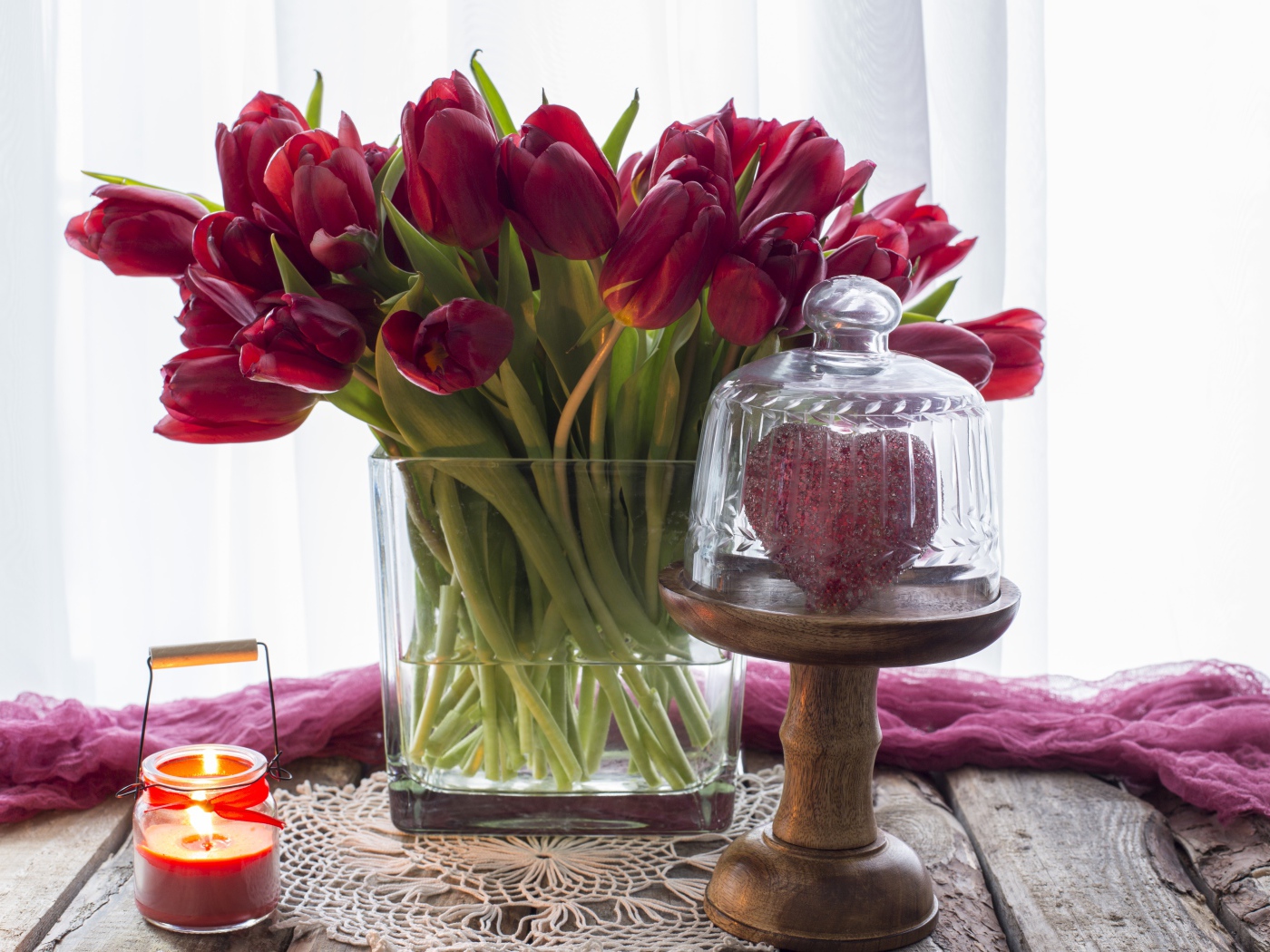 Букет красных тюльпанов в стеклянной вазе на столе со свечой и сердцем 