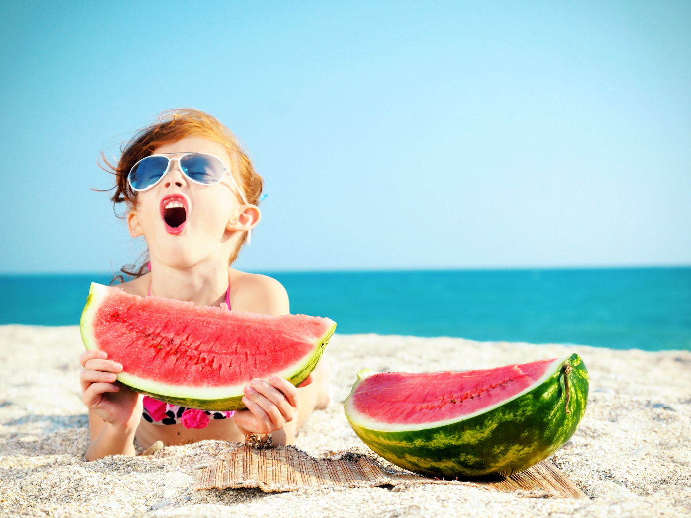 Маленькая девочка ест арбуз на песке у моря