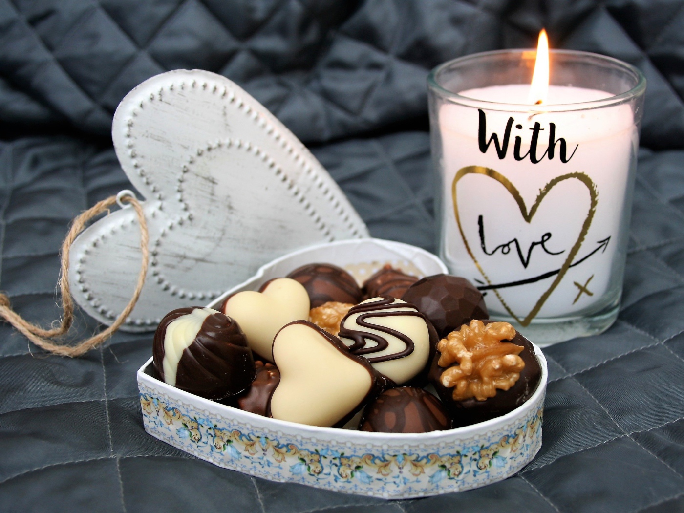 Шоколадные конфеты в коробке в форме сердца на кровати с зажженной свечой 