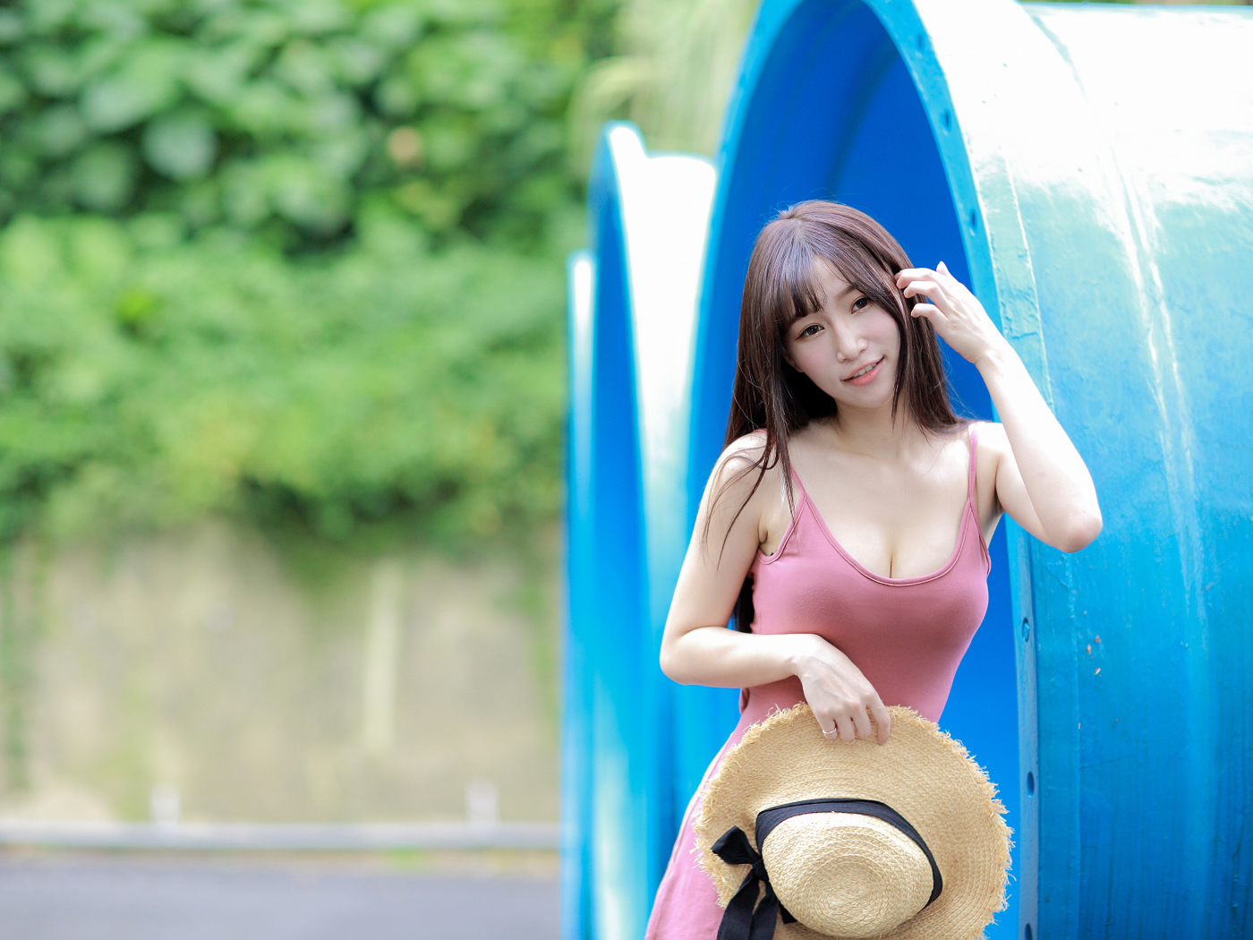 Стройная азиатка в платье со шляпой в руке стоит у синих  труб 