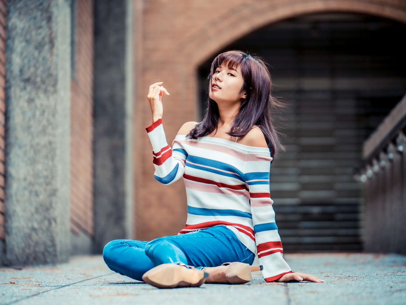 Девушка азиатка в джинсах и полосатом свитере сидит на асфальте