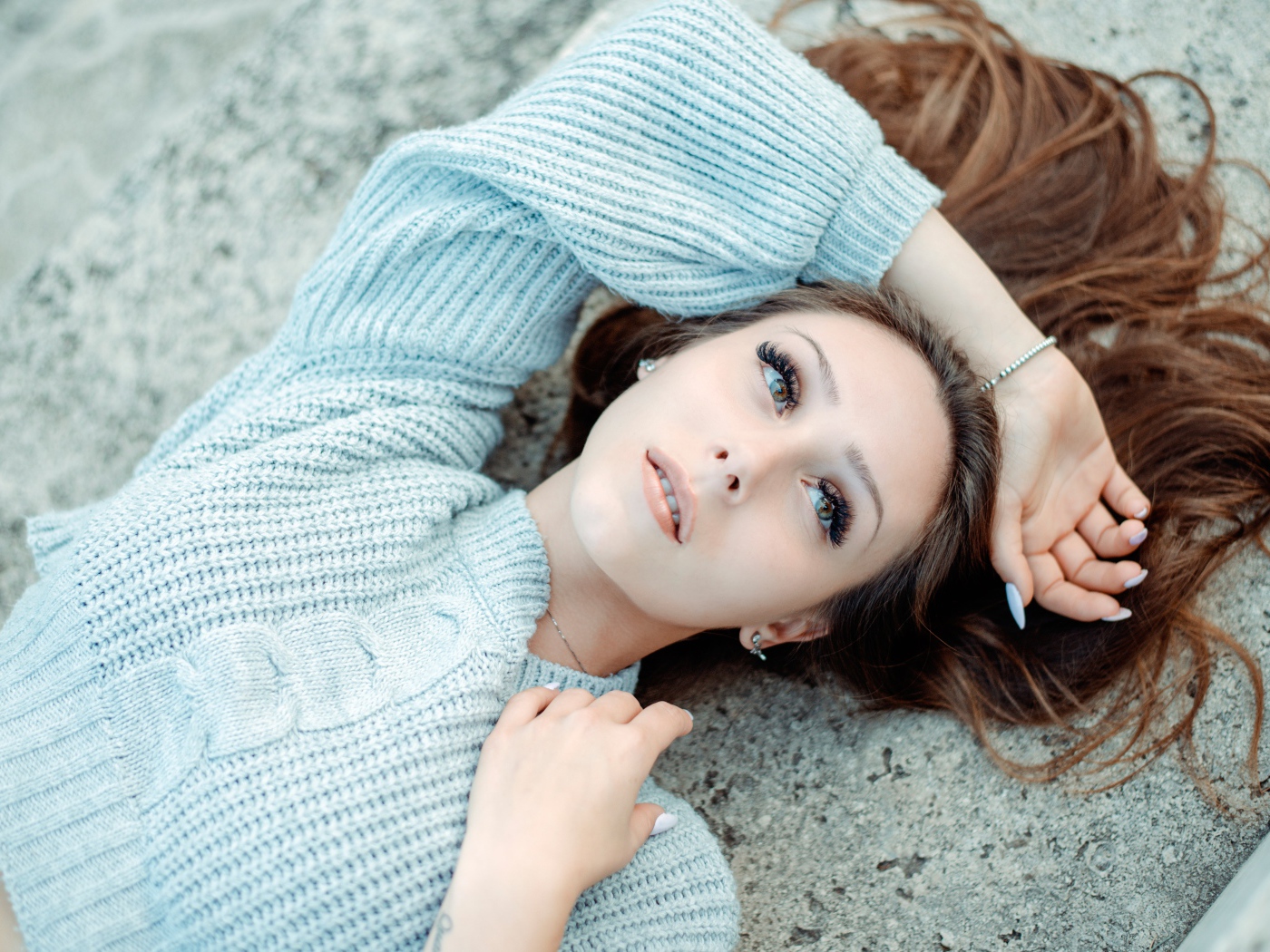 Длинноволосая девушка в голубом свитере лежит на земле 