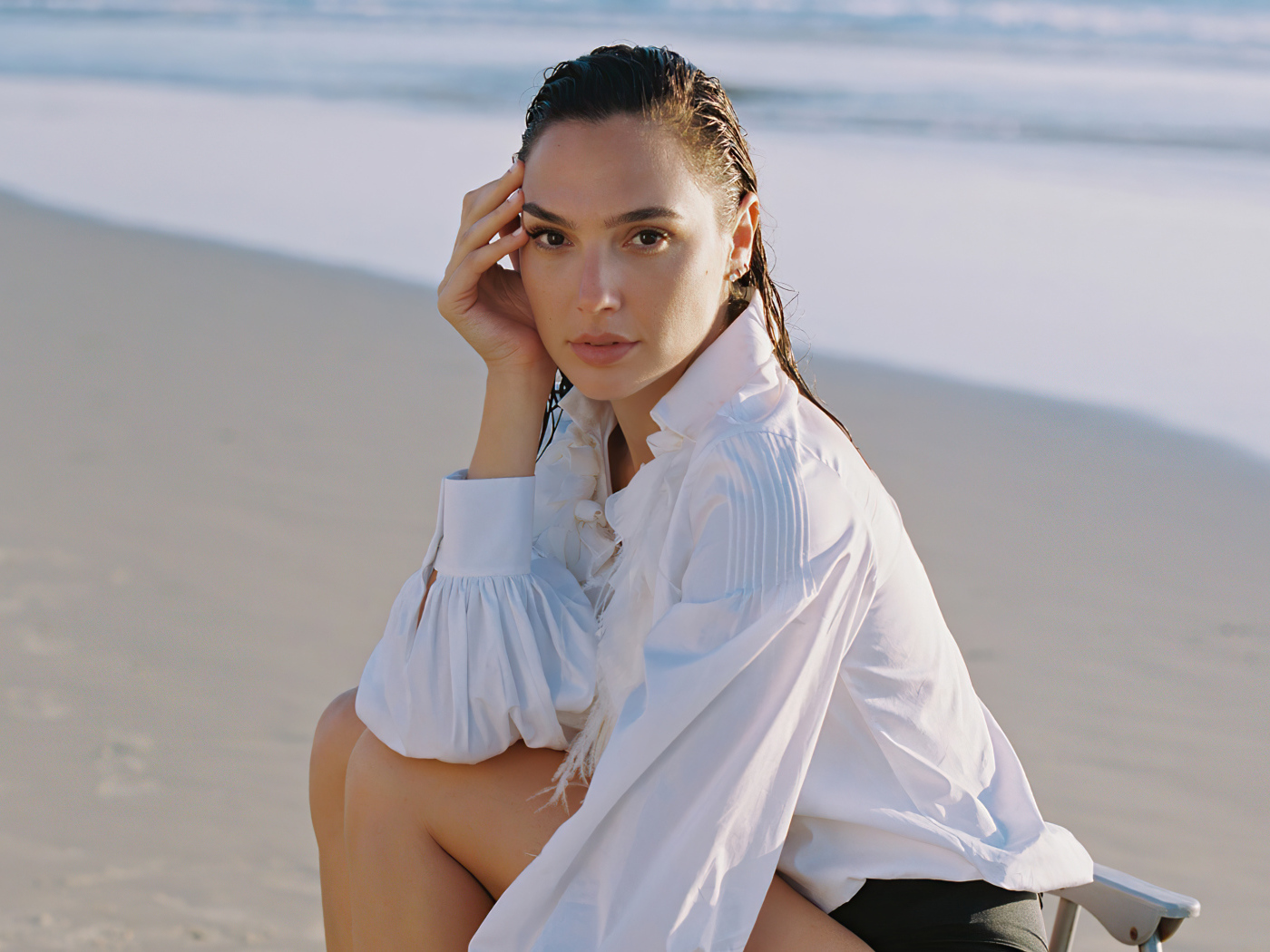 Актриса Галь Гадот в белой рубашке сидит на песке