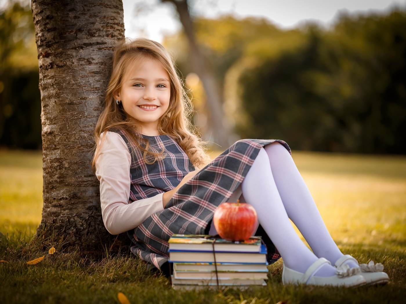 Маленькая девочка с книгами и яблоком сидит под деревом 