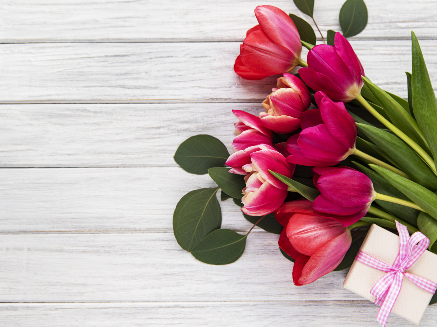 Букет розовых тюльпанов с подарком и зелеными листьями на белом столе