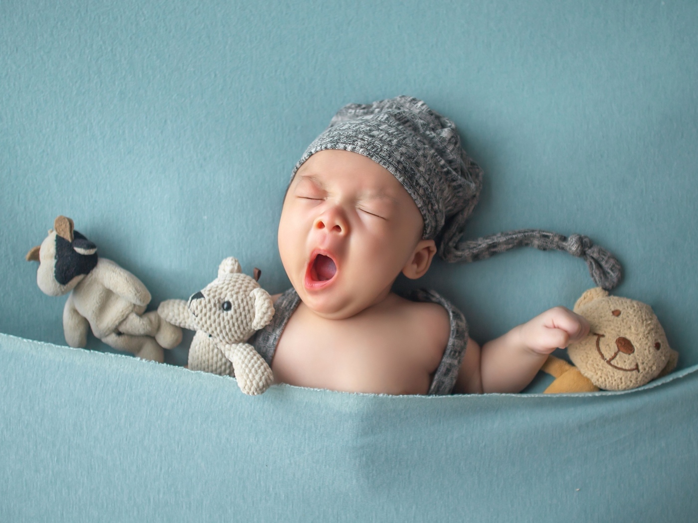 Зевающий малыш с игрушками под серым покрывалом