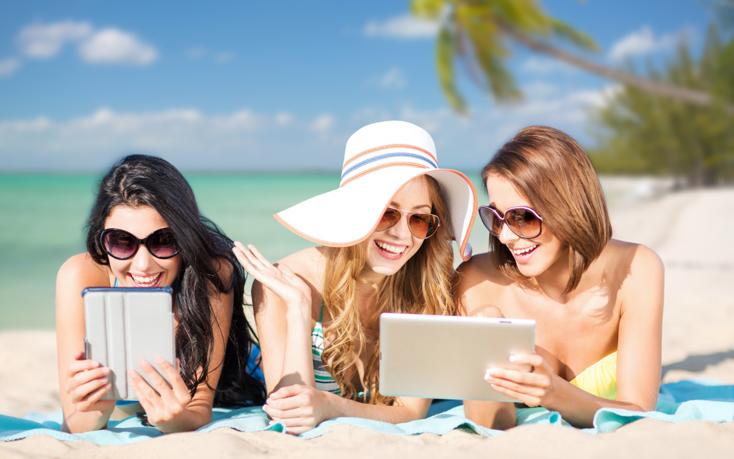 Три улыбающиеся девушки загорают на пляже с планшетами в руках