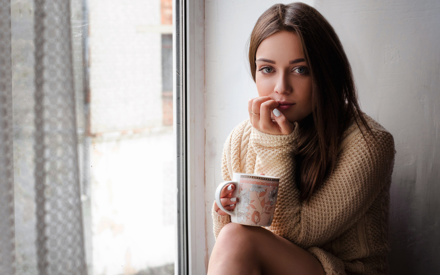 Молодая девушка в свитере сидит с чашкой на подоконнике