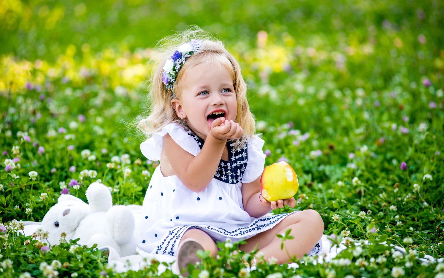 Маленькая улыбающаяся девочка с яблоком сидит на зеленой траве