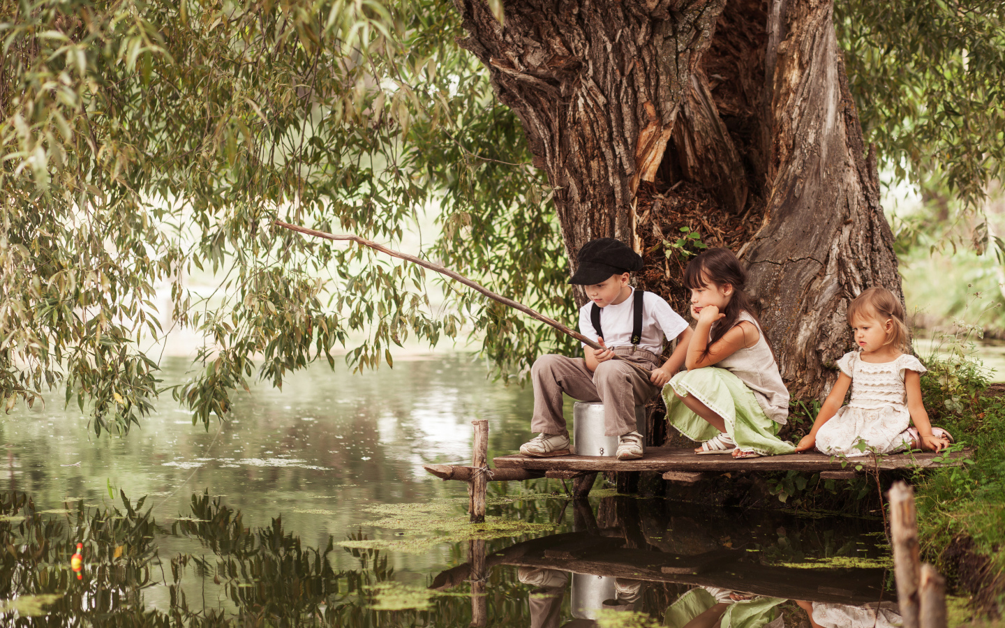 Две маленькие девочки и мальчик сидят на берегу пруда с удочкой