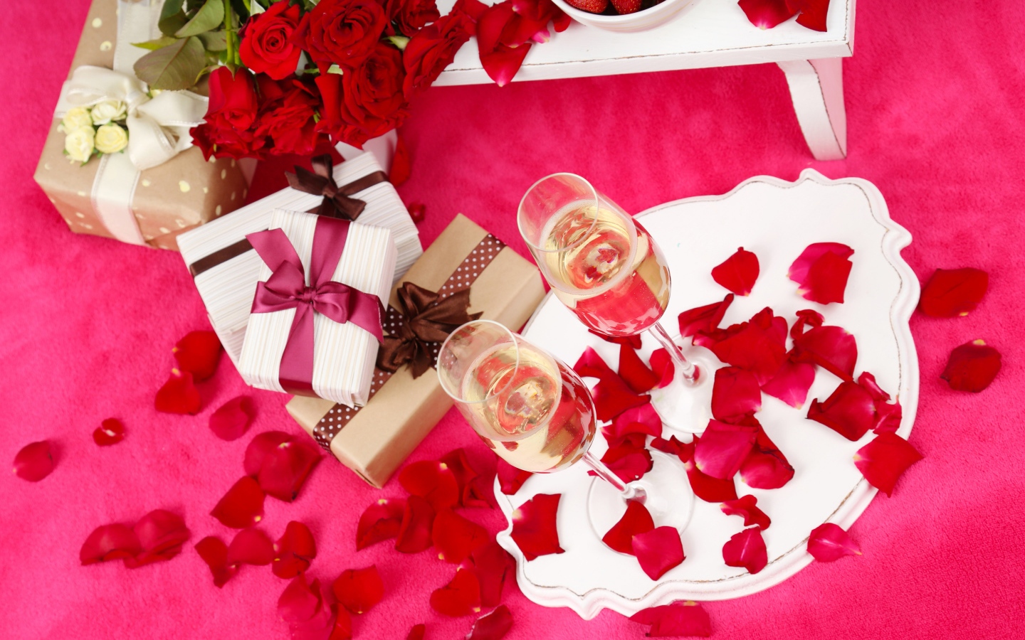 Два фужера с шампанским на красном покрывале с лепестками, подарками и букетом роз для любимой