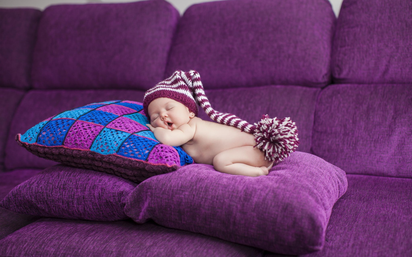 Маленький ребенок в большой шапке спит на фиолетовых подушках