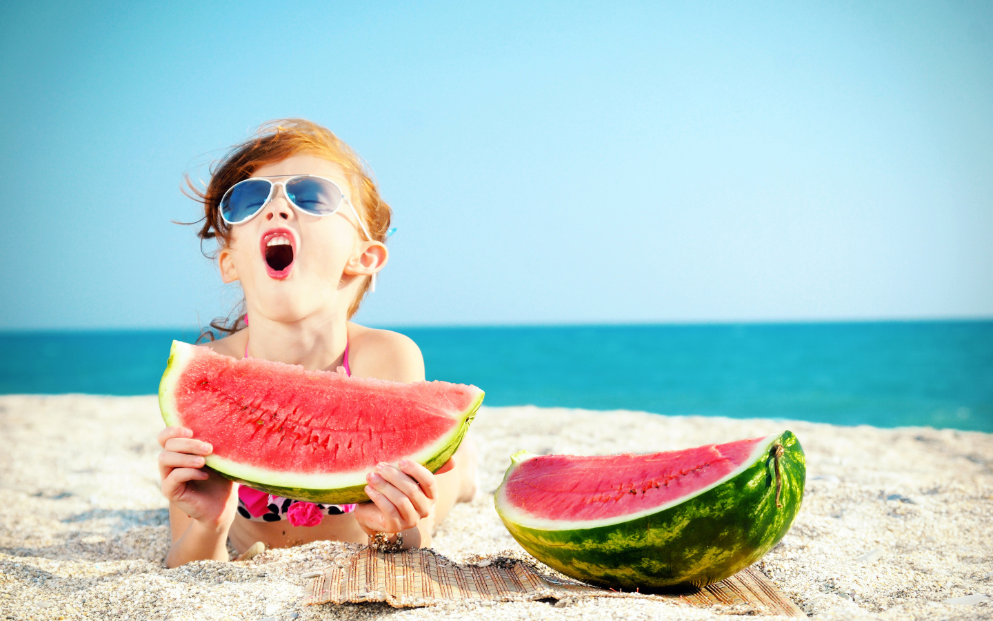 Маленькая девочка ест арбуз на песке у моря