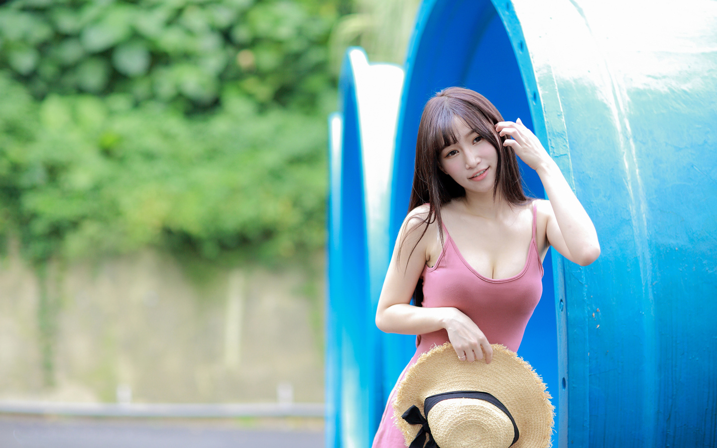 Стройная азиатка в платье со шляпой в руке стоит у синих  труб 