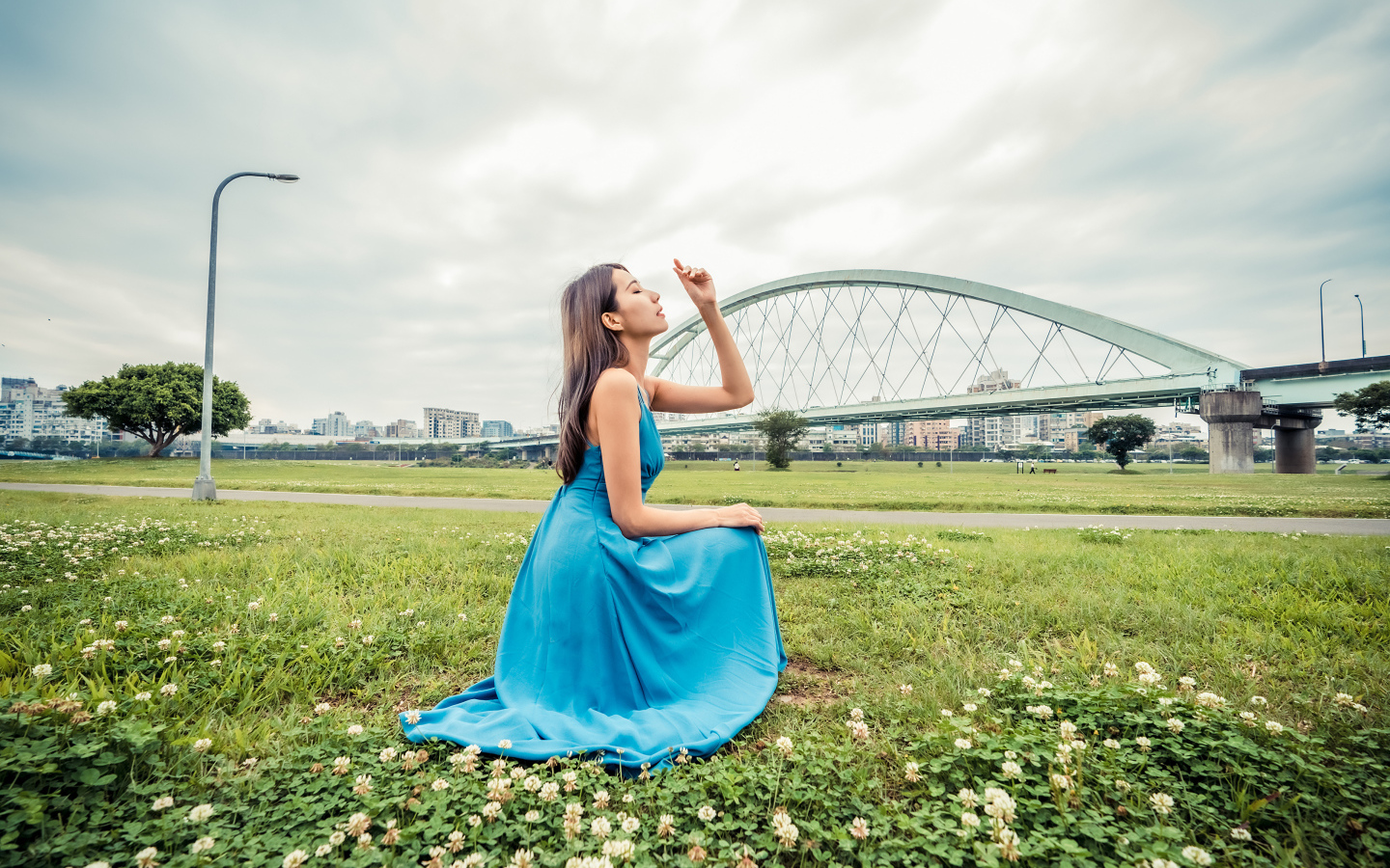 Красивая девушка азиатка в голубом платье  сидит на траве на фоне моста