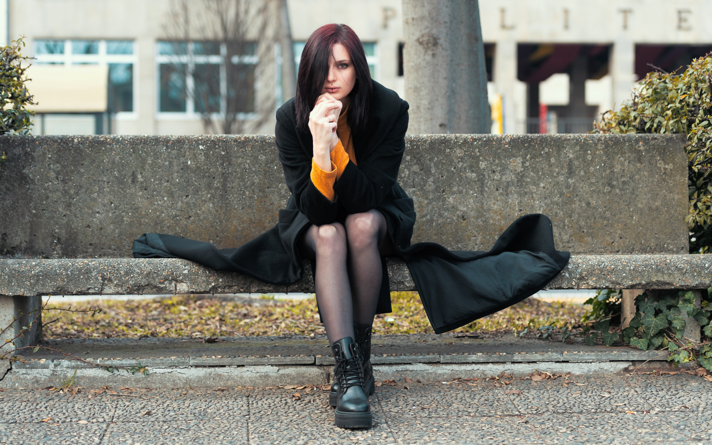 Девушка в черном пальто сидит на бетонной лавке 