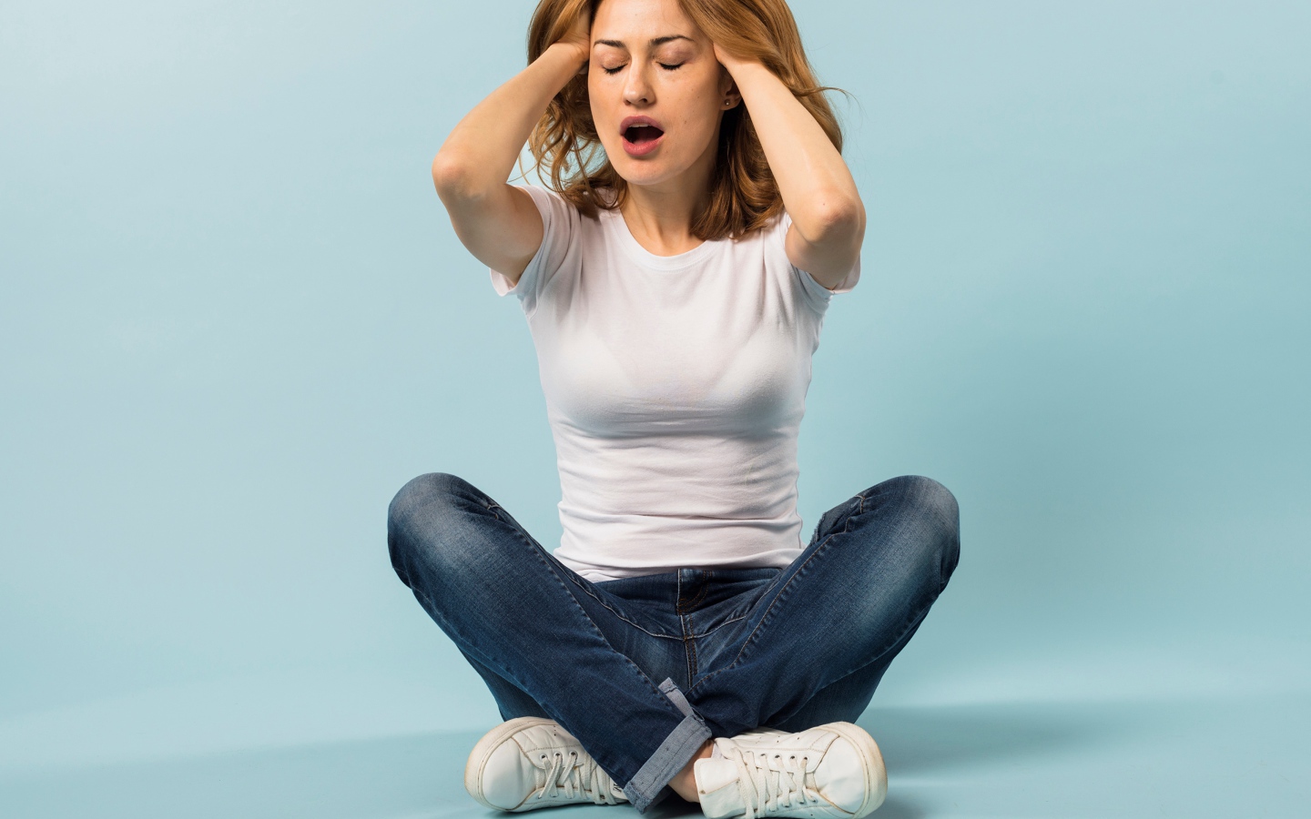 Девушка с руками в волосах сидит в джинсах на голубом фоне