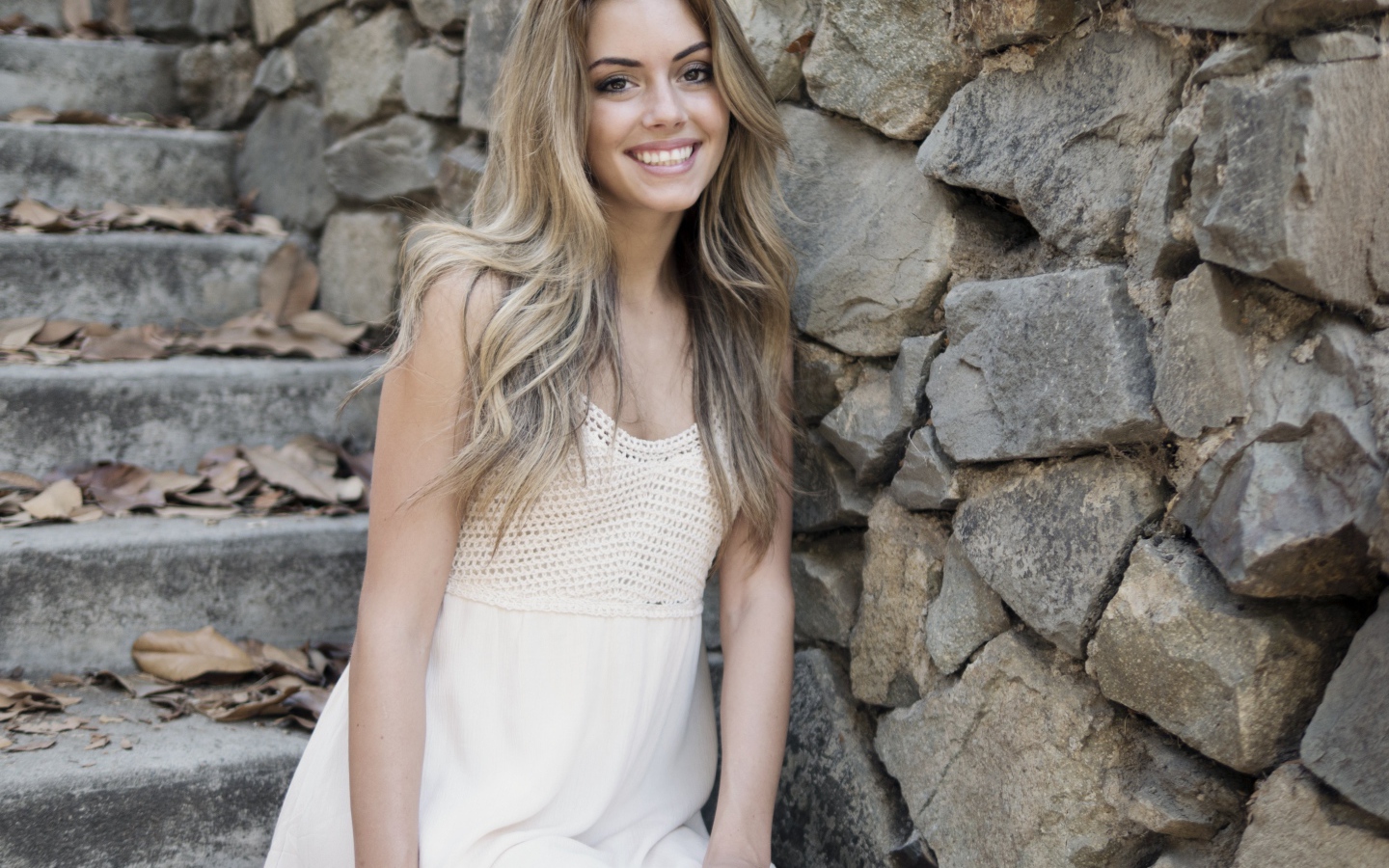 Улыбающаяся  девушка в белом платье стоит на ступеньках у стены